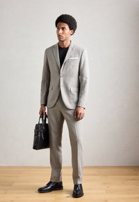Hombre con traje gris y camisa negra sosteniendo un maletín de cuero negro, de pie sobre un suelo de madera contra una pared clara y lisa.