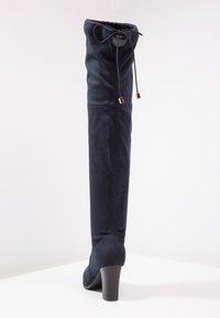 Bota de ante azul marino por encima de la rodilla con tacón cónico, con parte superior fruncida y cordón ajustable con detalles dorados para un ajuste seguro.