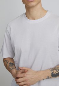 Witte katoenen T-shirt met een ronde hals en korte mouwen, met een gladde textuur en zichtbare tatoeages op de onderarmen.