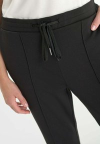 Close-up van zwarte broek met een trekkoord in de taille, gedragen door een persoon in een witte top, met de handen bijna bij de heupen.