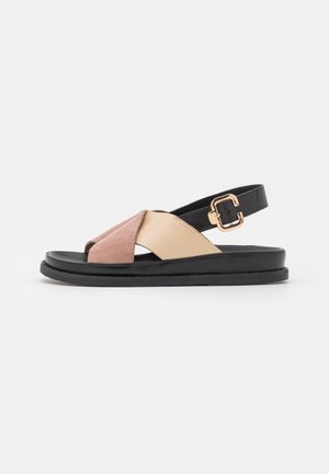 Flad sandal med sort sål, beige og pink ruskind krydset remme, sort ankelrem og roseguld spænde på hvid baggrund.