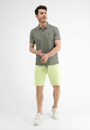 Mann trägt olivgrünes Poloshirt, hellgelbe Shorts mit Kordelzug und weiße Turnschuhe, steht vor einem schlichten weißen Hintergrund.