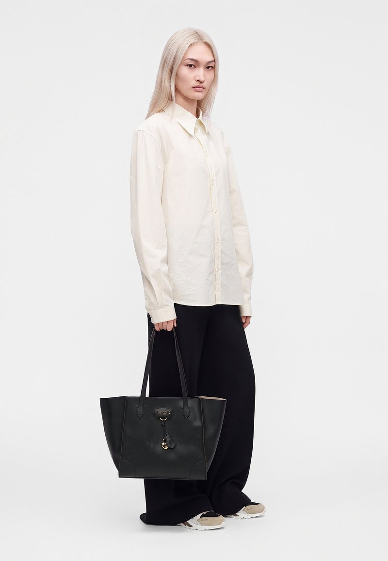 Femme aux longs cheveux blonds portant une chemise crème boutonnée et un pantalon noir, tenant un grand sac cabas noir et des baskets beige.