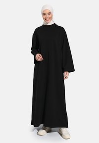 Black maxi dress with long sleeves and a high neckline, made of a soft fabric. The model wears white sneakers and a light-colored hijab.
