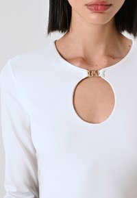 Maglietta bianca a maniche lunghe con scollatura a oblò e un accento decorativo dorato sul colletto. Tessuto liscio e aderente senza motivi visibili.