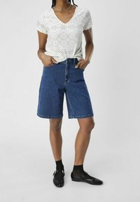 Weißes gehäkeltes Kurzarmoberteil mit V-Ausschnitt, kombiniert mit dunkelblauen Denim-Bermudashorts und schwarzen flachen Schuhen.
