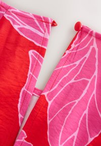Close-up of a red and pink fabric with white leaf patterns and a red fabric-covered button at the edge on a white background.