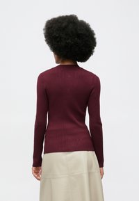 Pessoa com cabelo encaracolado preto, usando uma blusa justa de manga longa em tom bordeaux, e uma saia bege clara, de costas para a câmera.