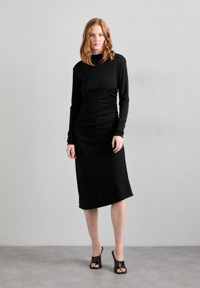 Robe noire à manches longues avec texture froncée, coupe ajustée, longueur genou, associée à des chaussures à talons ouverts noires sur un fond gris.