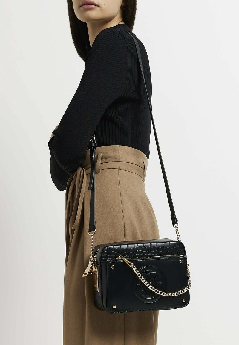 Svart crossbody-väska med krokodilmönstrad textur och detaljer i guld. Har en avtagbar kedjerem och logotypdetalj på framsidan.