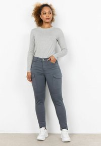 Šedé tričko s dlouhým rukávem v kombinaci se šedými jogger kalhotami s kapsami, slim fit a hladkou texturou. Boty jsou bílé sportovního stylu.