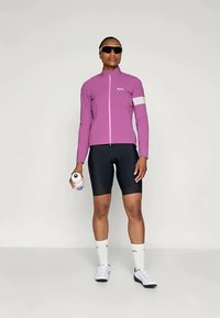 Athlète portant une veste violette, un short de cyclisme noir, des chaussettes blanches, des lunettes de soleil et tenant une bouteille d'eau blanche, se tenant devant un fond uni.