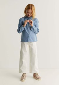Fată îmbrăcată cu o cămașă albastră cu nasturi și pantaloni albi largi, uitându-se în jos și ajustându-și degetele, stand pe un fundal simplu.