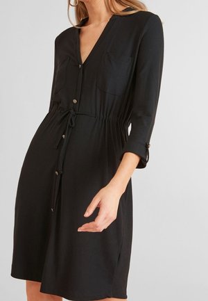 Robe noire mi-longue avec fermeture à boutons devant, taille avec cordon, poches poitrine et manches trois-quarts sur une personne devant un fond uni.