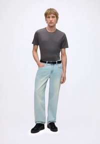 Tânăr purtând un tricou fitted gri închis, blugi albastru deschis cu o curea neagră și pantofi negri, stând în picioare pe un fundal alb simplu.