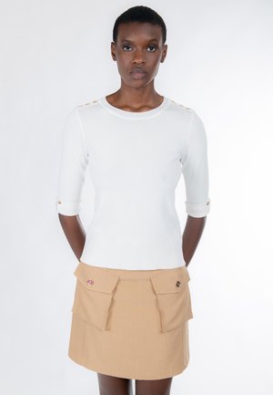 Donna che indossa una maglia bianca a maniche tre quarti con bottoni sulle spalle e una gonna beige con grandi tasche frontali, in piedi con le mani dietro la schiena.