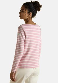 Kobieta z kręconymi włosami w kokach, ubrana w różową i białą w paski koszulę z długim rękawem oraz prawie białe spodnie, stojąca bokiem z lekko odwróconym plecami.