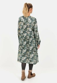 Kvinde med blond hestehale iført en lang grøn blomsterkjole, sort leggings, beige sokker og brune ankelstøvler, vendt væk.