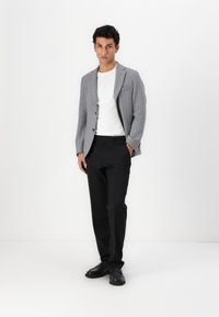 Blazer grigio con righe sottili e una singola tasca sul petto, indossato sopra una t-shirt bianca e pantaloni neri, abbinato a scarpe nere.