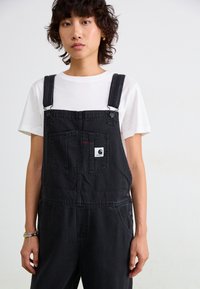 Schwarze Denim-Latzhose mit verstellbaren Trägern, Fronttasche mit Logo, kombiniert mit einem weißen T-Shirt. Glatte Textur, einfaches Design.