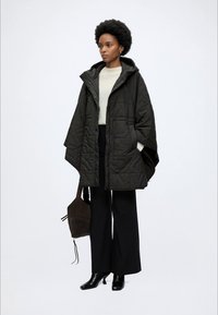 Γυναίκα με μαύρη καπιτονέ κάπα με κουκούλα, λευκό πουλόβερ, μαύρο παντελόνι με φαρδιά μπατζάκια, μαύρα μποτάκια μέχρι τον αστράγαλο, κρατώντας καφέ τσάντα στο πλάι.