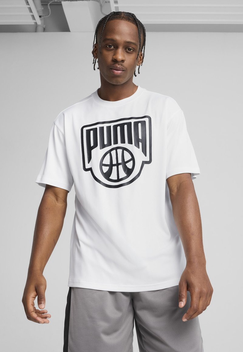 Młody mężczyzna z warkoczami, noszący białą koszulkę z logo Puma do koszykówki i szare szorty sportowe, na jednolitym tle.
