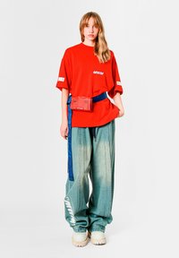 Camiseta roja de gran tamaño con estampado "MWM", combinada con vaqueros holgados azul claro. Completa el conjunto una pequeña riñonera roja y un cinturón azul. Zapatos blancos gruesos.