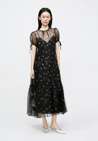 Rochie neagră cu imprimeu floral, mâneci bufante transparente, design în straturi și textură ușoară. Poartă pantofi comozi verzi pal cu vârf ascuțit și o geantă neagră.