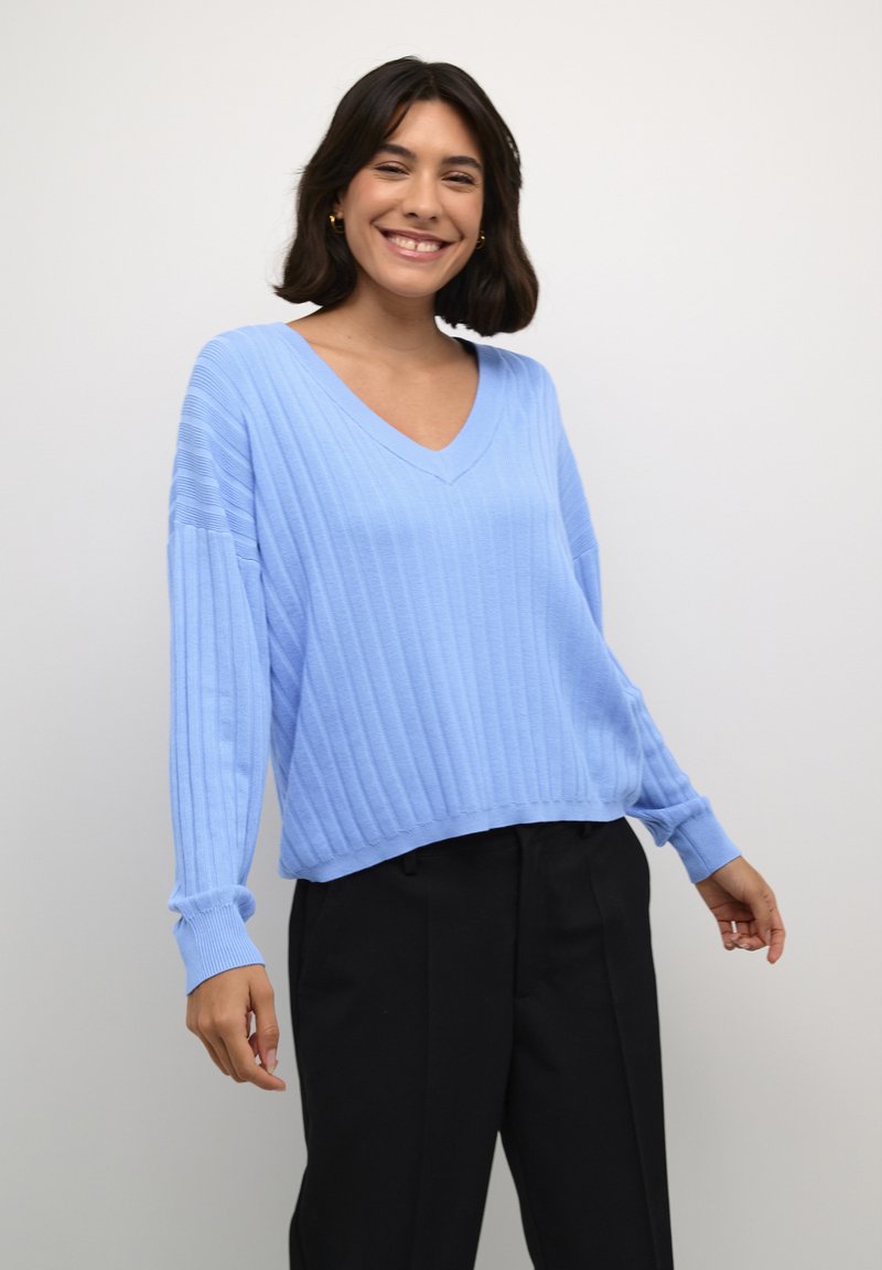 Šviesiai mėlynas lygaus rašto megztinis su V formos iškirpte ir ilgomis rankovėmis; tekstūruotas audinys su laisvu siluetu, derinamas su juodomis kelnėmis.