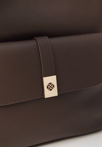 Sac en cuir marron à la texture lisse, doté d'un fermoir doré avec un design découpé complexe. Détails cousus sur les bords.
