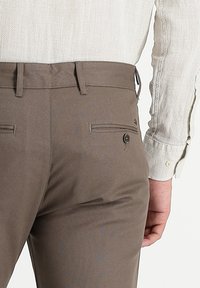 Man wearing brown tailored trousers and a textured beige long-sleeve shirt, hand resting by side, shown from the waist down.