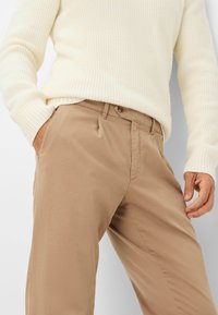 Chinos beige con una texture liscia, caratterizzati da un davanti piatto, tasche laterali e una vita con bottone. Abbinati a un maglione in maglia crema.