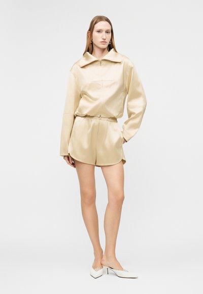 Γυναίκα που φοράει μακρυμάνικη μπλούζα από μπεζ σατέν με φερμουάρ και ταιριαστό σορτς, στέκεται με το ένα χέρι στην τσέπη και λευκά mule με τακούνι.