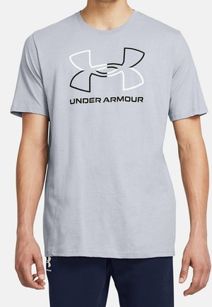 Mann trägt ein hellgraues Under Armour T-Shirt mit großem schwarz-weißem Logo und Text auf der Brust, kombiniert mit dunkler Hose.