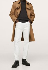 Brauner Trenchcoat über einem dunklen Strickpullover, kombiniert mit weißen Hosen und schwarzen Knöchelstiefeln. Glatte Textur und lässige Passform.