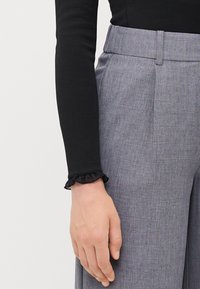 Black ribbed long-sleeve top with a ruffled sheer cuff, paired with light grey tailored trousers featuring a subtle textured pattern and pleats.
