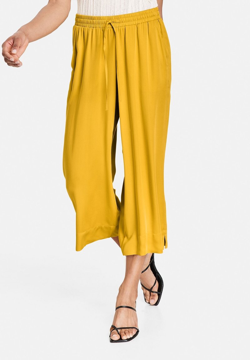 Żółte spodnie szerokie z elastycznym pasem i sznurkiem. Gładki materiał z bocznymi rozcięciami na dole. Połączone z czarnymi sandałami na paski.