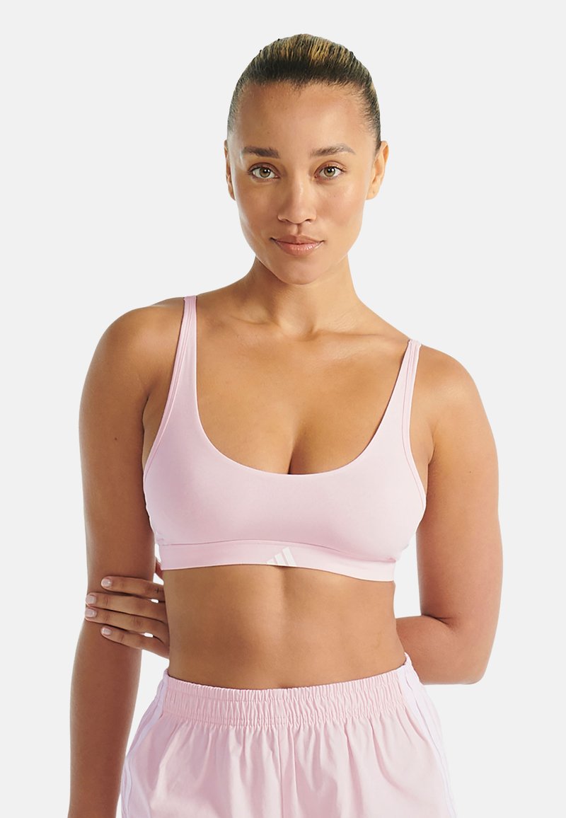 Kobieta z związanymi włosami, ubrana w jasnoróżowy stanik sportowy i dopasowane szorty, stoi z jedną ręką skrzyżowaną za plecami.