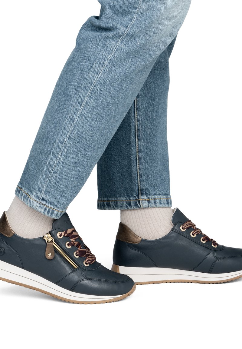 Personne portant un jean bleu clair, des chaussettes crème et des baskets bleu foncé avec des détails marron, une fermeture éclair dorée et des semelles blanches, avançant d'un pas.