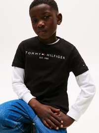 Μαύρο κοντομάνικο t-shirt με λευκό λογότυπο "TOMMY HILFIGER EST. 1985", τοποθετημένο πάνω από μια λευκή μακρυμάνικη μπλούζα. Denim jeans σε μπλε χρώμα.