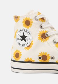 Bílé vysoké tenisky Converse s motivem slunečnic, kovovými očky, bílými šnůrkami a kulatou černobílou nášivkou na kotníku.
