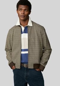 Checkered brown bomber jacket with a ribbed dark brown hem, layered over a blue and white striped polo shirt, with a textured finish.