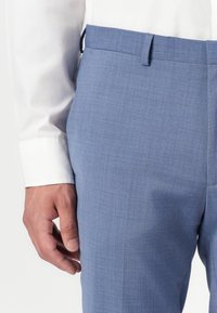 Világoskék, testre szabott nadrág, texturált anyagból, lapos elejű, övhurkokkal és slim fit szabással, egy fehér inggel viselve.