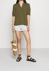 Blusa verde oliva con bottoni e maniche corte, abbinata a pantaloni corti in denim azzurro, sandali neri e una borsa intrecciata con pattern a righe.