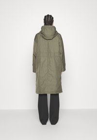 Cappotto lungo verde oliva con cappuccio, dalla vestibilità dritta, realizzato in materiale liscio. Presenta tasche laterali e polsini regolabili.