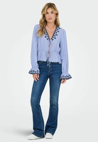 Lichtblauwe blouse met lange mouwen en een diepe V-hals, met een geplooide taille en zwarte bloemenborduursels langs de halslijn en manchetten, gecombineerd met uitlopende denim jeans.