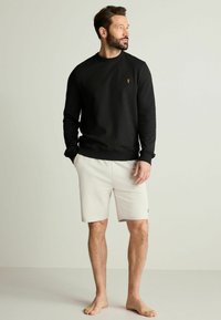 Sudadera negra de manga larga con cuello redondo, que presenta un pequeño logo. Combinada con pantalones cortos de un tono beige claro, exhibida en un modelo descalzo.