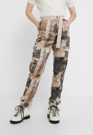 Osoba ubrana w beżowe spodnie bojówki w wzory z paskiem, biały prążkowany krótki rękaw i białe sznurowane botki, stojąca z rękami w kieszeniach.