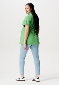 Vrouw met lange vlechten, gekleed in een groen t-shirt met korte mouwen, lichte spijkerbroek en witte sneakers, staand en over haar schouder kijkend.