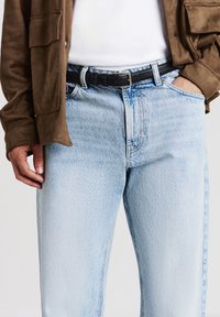 Vista ravvicinata di una persona che indossa jeans azzurri chiari con cintura nera, camicia bianca e giacca marrone; una mano nelle tasche, l'altra rilassata lungo il corpo.